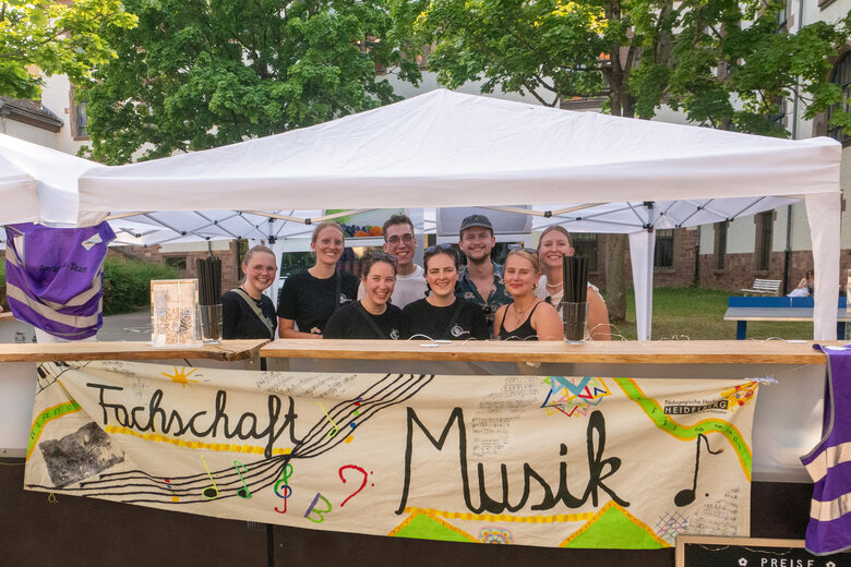8 Studierende der Fachschaft Musik stehen bei einer Veranstaltung im Innenhof der PH hinter der Bar und lachen in die Kamera.
