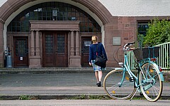 Das Bild zeigt eine junge Frau von hinten. Sie läuft auf das Altbauportal der Hochschule zu. Vorne rechts sieht man ein Fahrrad.