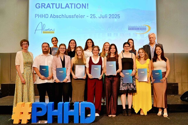 Zu sehen sind Alumni, die auf einer Bühne stehen und ihre Bestehensbriefe in PHHD-Mappen hochhalten. Gemeinsam mit Rektorin Vach & Prorektor Dammer (a.D.), welche ihnen die Mappen überreicht haben.