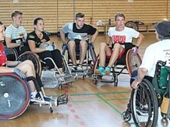 Eine Gruppe rollstuhlfahrender Sportler:innen sitzt im Halbkreis vor einer weiteren Person. Sie befinden sich in einer Sporthalle.