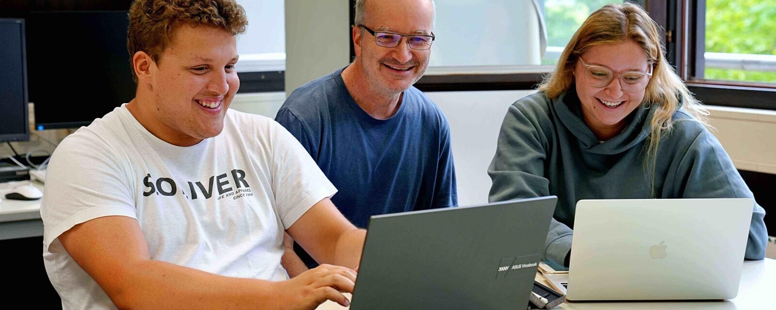 Ein Student, ein Dozent und eine Studentin arbeiten gemeinsam an Laptops. Sie alle schauen freundlich.