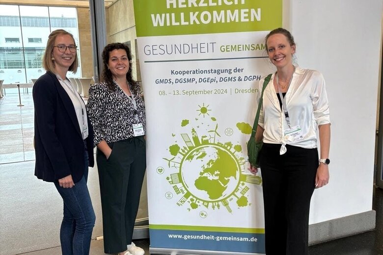 Auf dem Bild sind drei Personen zu sehen, die vor einem Kongressposter stehen, welches sich um die Themen Gesundheit und Präventionsförderung handelt.