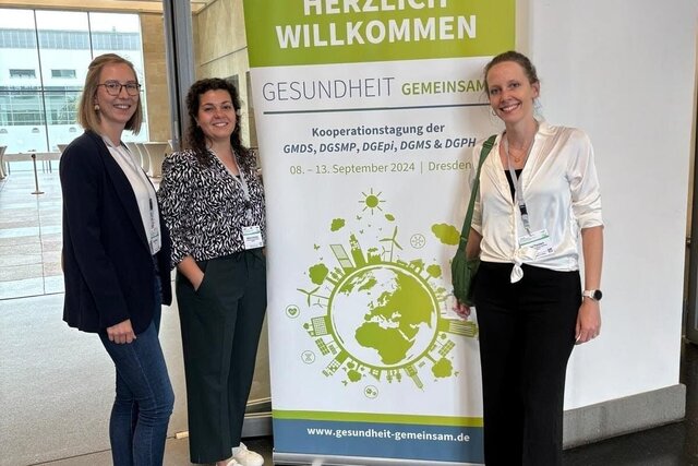 Auf dem Bild sind drei Personen zu sehen, die vor einem Kongressposter stehen, welches sich um die Themen Gesundheit und Präventionsförderung handelt.