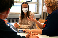 Studentin sitzt mit Karin Terfloth an einem Tisch. Terfloth spricht deutlich mit den Händen. Vorne links sieht man eine Person von hinten. Alle tragen Corona-bedingt einen Mund-Nase-Schutz.