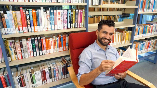 Junger Mann sitzt auf einem Stuhl in der Bibliothek, welche sich direkt am Campus der Pädagogischen Hochschule Heidelberg befindet. Hinter ihm eine Bücherregalwand.