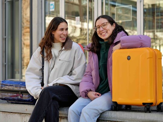 Zwei Studierende sitzen mit einem Koffer vor einem Gebäude der PHHD.