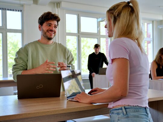 Ein Mann spricht deutlich mit den Händen. Er schaut zu einer Frau, die rechts im Bild steht. Im Hintergrund arbeiten zwei Personen an Stehtischen.