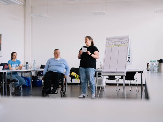 Das Foto zeigt drei Menschen neben einer Flipchart. Diese halten gerade einen Vortrag.
