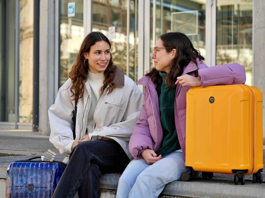 Zwei Studentinnen der PH Heidelberg sitzen mit gepackten Koffern auf einer Stufe vor dem Eingang eines Gebäudes.