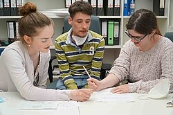 Das Bild zeigt drei Menschen, die über einen Schreibtsich gebeugt sitzen und arbeiten. drei Studierende