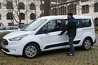 Auf dem Bild ist ein weißes Auto zu sehen. Klemens steht an der Tür und greift zu dem Griff. Das Auto steht im Innenhof der alten PH.