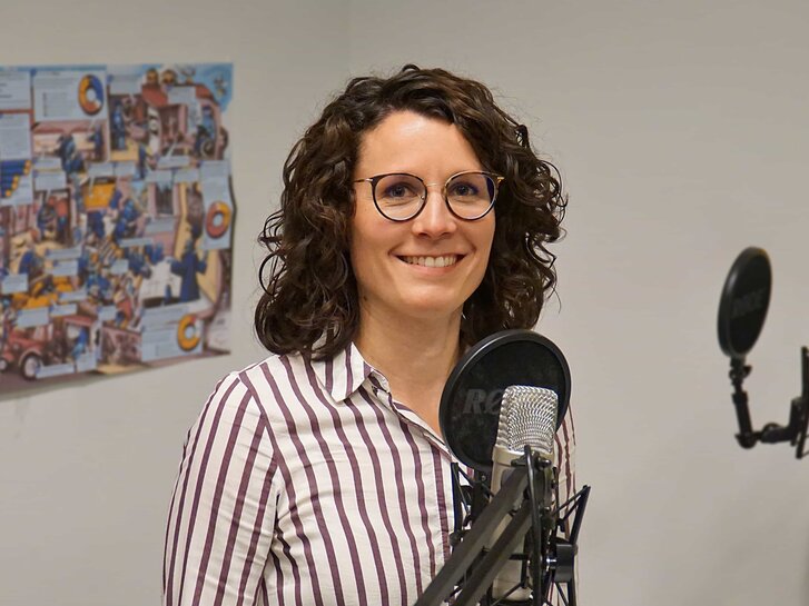 Das Bild zeigt Christina Mechler im Podcast-Studio. Sie steht vor zwei Mikrofonen und lächelt in die Kamera.