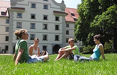 Das Bild zeigt vordergründig vier Studierende, die gemeinsam auf einer grünen Wiese sitzen. Im Hintergrund ist ein Gebäude der Pädagogischen Hochschule Weingarten zu sehen.