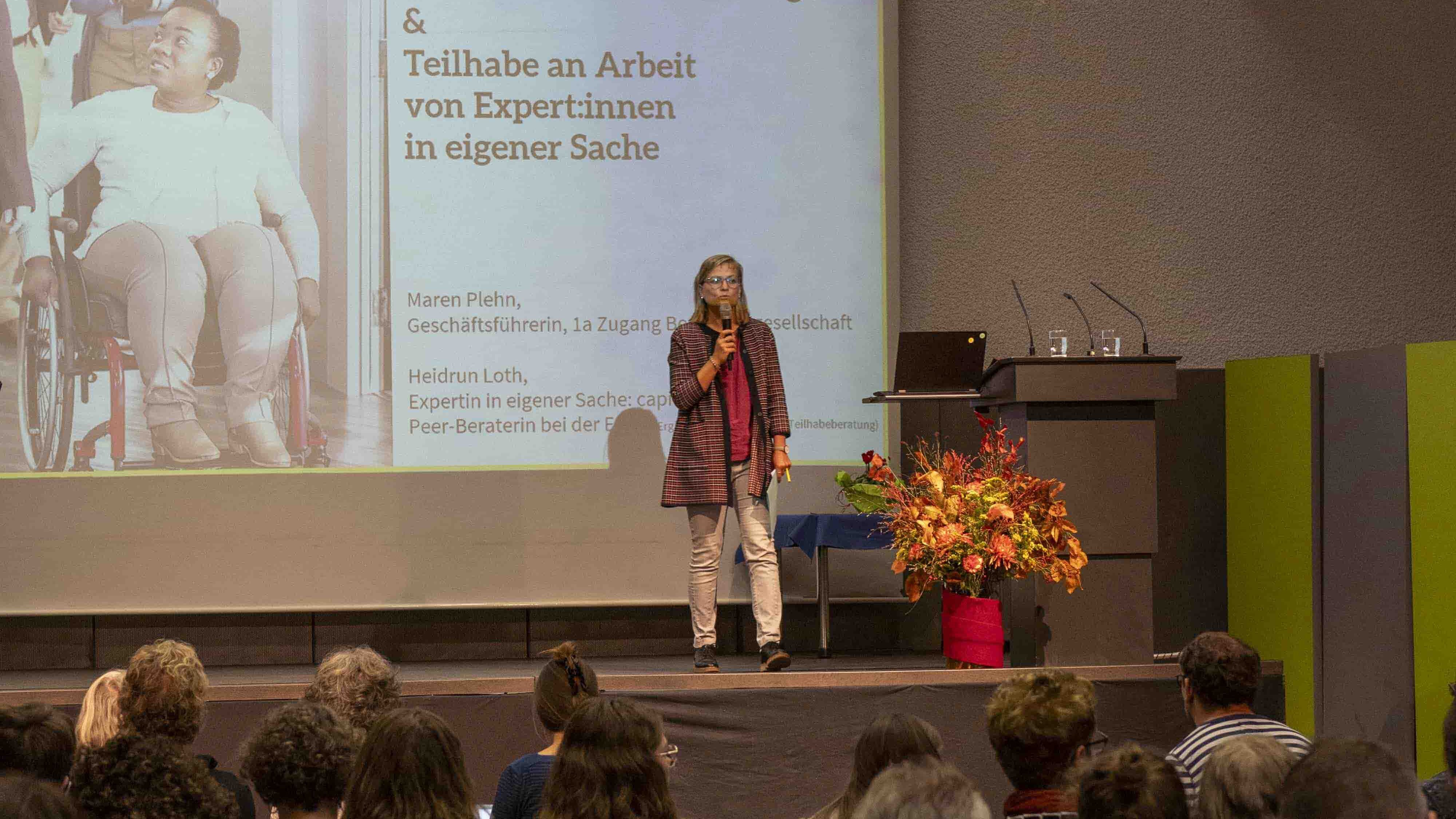 Eine Frau steht auf der Bühne in der Mehrzweckhalle der PH Heidelberg. Sie hält einen Vortrag.