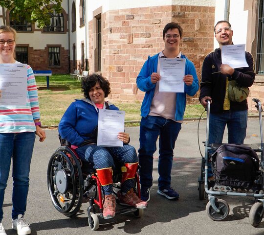 Gruppenbild der Qualifizierungsteilnehmenden: Lilly Lorenz, Maya Silva, Cedric Rueter und Mehmet Can Friedel sind im Innenhof der PH Heidelberg und halten ihre Zertifikate zur bestandenen Modulprüfung in den Händen. Alle lächeln in die Kamera.
