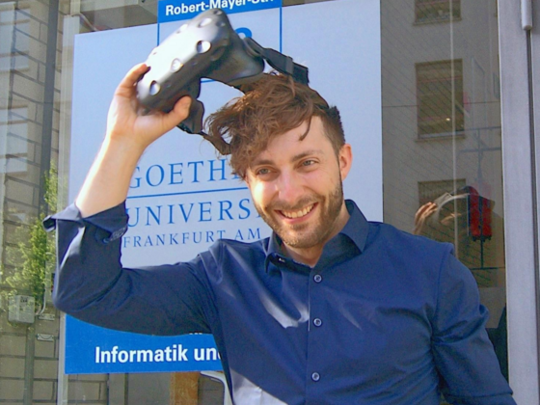 Prof. Dr. Andreas Dengel zieht sich eine VR-Brille vom Kopf, im Hintergrund ein Schild der Goethe-Universität Frankfurt.