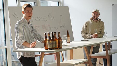 Zu sehen sind die beiden Gründer von Heerlijk Bier an einem Tisch stehend. Vor ihnen einige Bierflaschen.