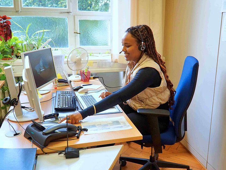 Eine Frau sitzt am Schreibtisch in ihrem Büro des SSC und beantwortet mit einem Headset die Fragen, die per Telefon gestellt werden.