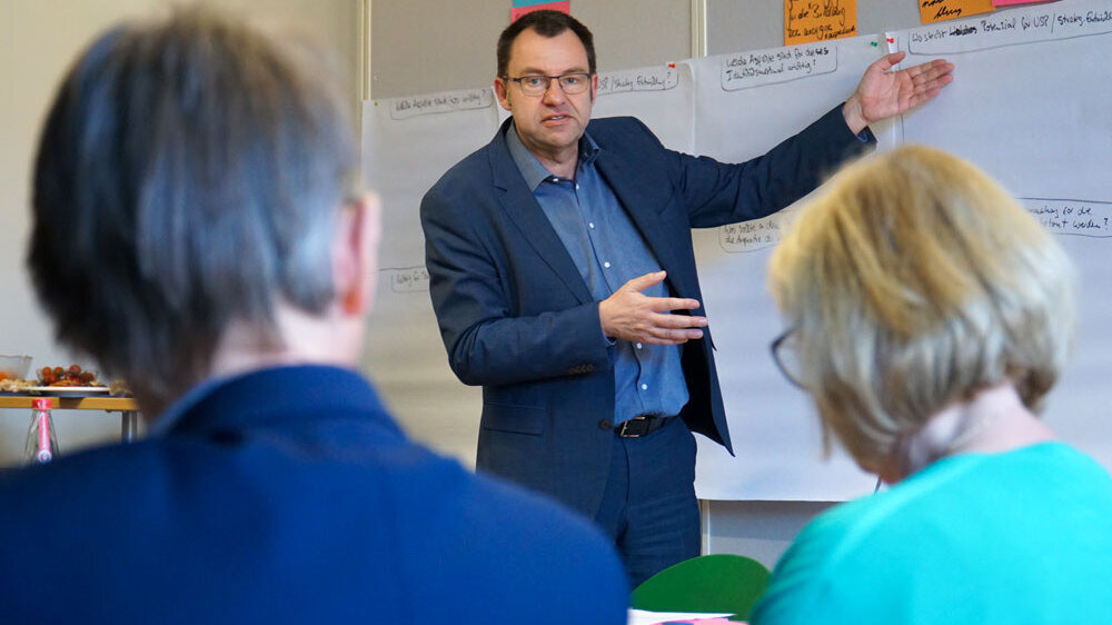 Frank Ziegele schaut direkt in die Kamera und spricht deutlich mit den Händen in die Richtung einer Pinnwand.