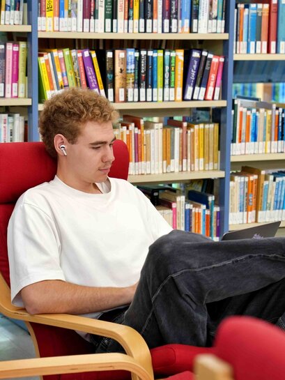 Zu sehen ist ein Student in einem Lesesessel. Hinter ihm steht ein Bücherregal.