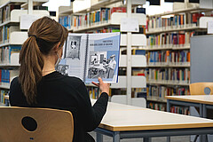 Auf dem Bild sieht man eine Frau von hinten. Sie sitzt in der Bibliothek der Hochschule und liest den Jahresbericht. 