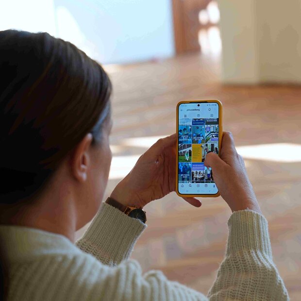 Eine Frau der Presse und Kommunikation sitzt auf der Treppe und blickt auf ihr Handy. Auf dem Handy sieht man die Instagram-Account der Hochschule.