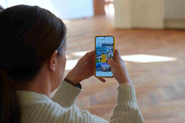 Eine Frau der Presse und Kommunikation sitzt auf der Treppe und blickt auf ihr Handy. Auf dem Handy sieht man die Instagram-Account der Hochschule.