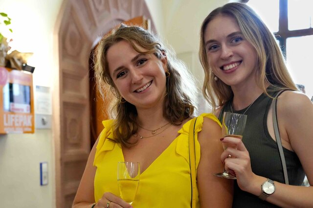 Zu sehen sind zwei Frauen mit einem Glas Sekt, die in die Kamera lachen.