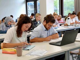 Studierende sitzen an Tischreihen und schreiben auf Tablets.