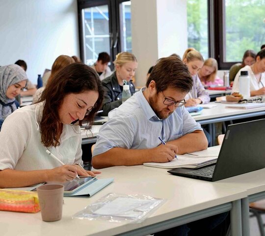 Studierende sitzen an Tischreihen und schreiben auf Tablets.