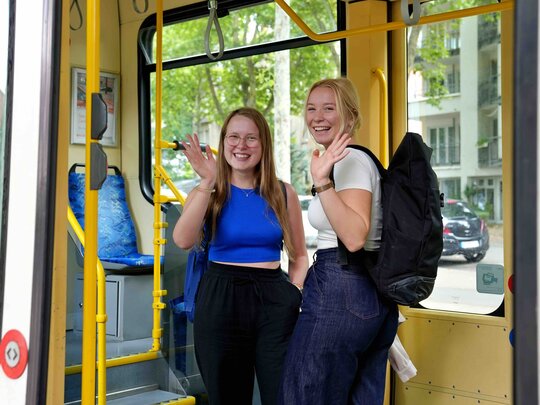Zwei Studierende stehen in einer Straßenbahn und winkeln fröhlich.