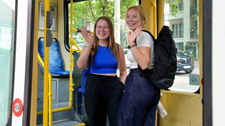 Zwei Studierende stehen in einer Straßenbahn und winkeln fröhlich.