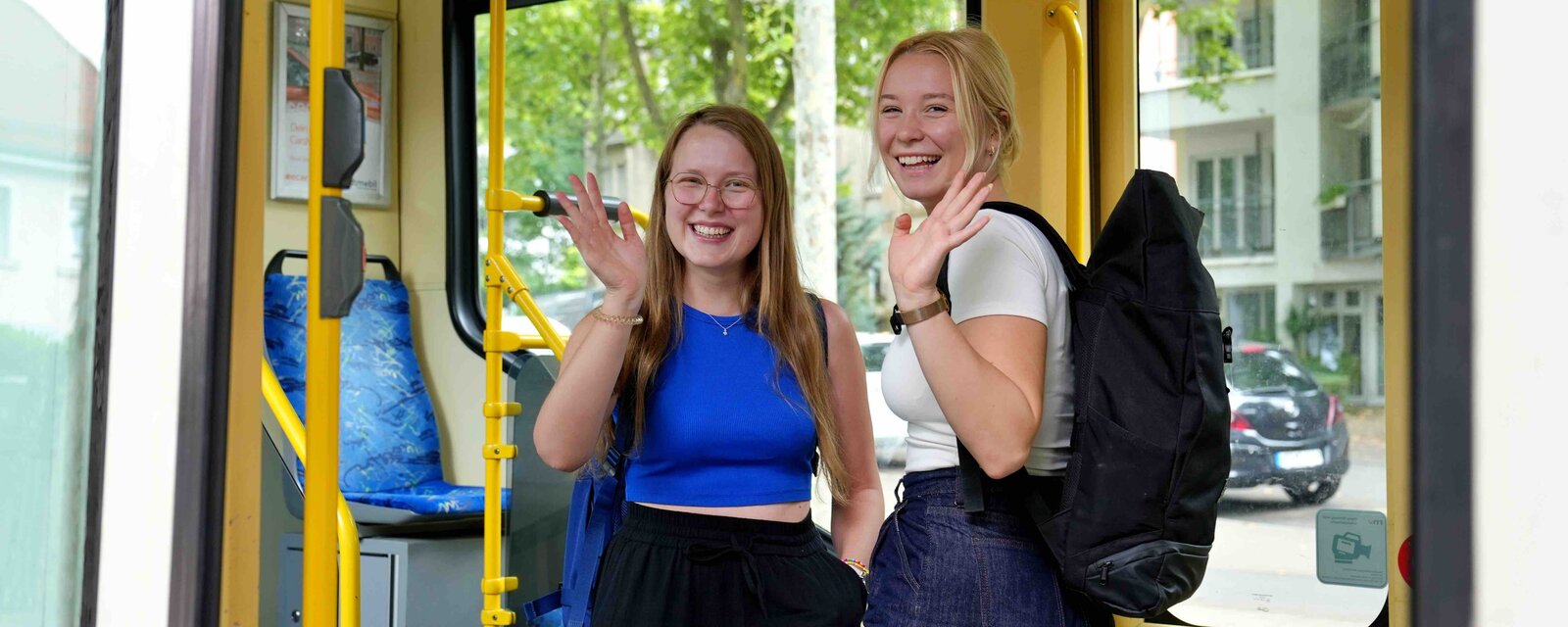 Zwei Studierende stehen in einer Straßenbahn und winkeln fröhlich.