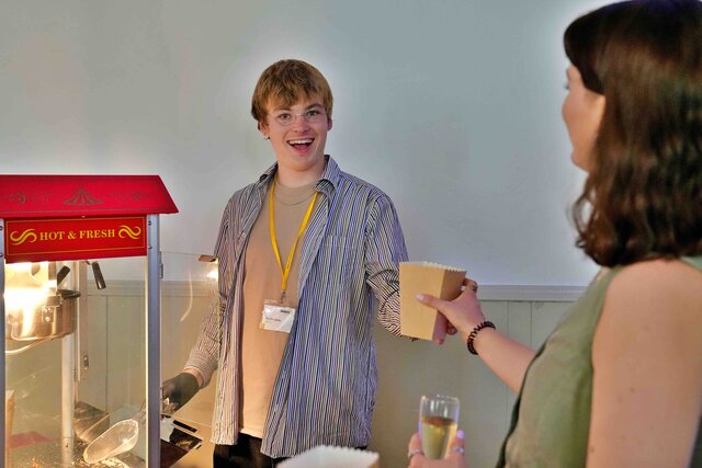 Zu sehen ist die studentische Hilfskraft Moritz an einer Popcorn-Maschine, wie er lachend Popcorn an eine Gästin ausgibt.