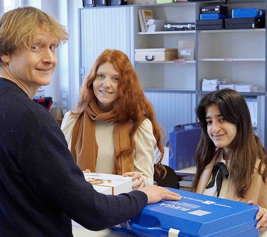 Drei Studenten stehen gemeinsam um einen blauen Koffer.