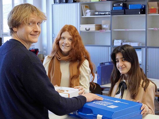 Drei Studenten stehen gemeinsam um einen blauen Koffer.