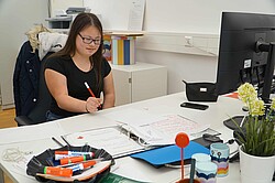 Das Foto zeigt eine junge Frau, die an einem Schreibtisch sitzt und mit dem Stift schreibt.