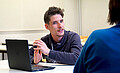 Michael Gänßmantel sitzt an einem Tisch, vor ihm ein Laptop. Er spricht mit einer Person, deren Rücken man sieht. Copyright: PH Heidelberg