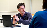 Das Bild zeigt Michael Gänßmantel. Er sitzt an einem Tisch, vor ihm ein Laptop. Er spricht mit einer Person, deren Rücken man sieht. Copyright: PH Heidelberg