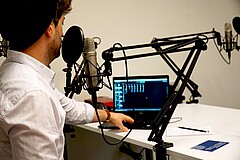 Das Bild zeigt einen jungen Mann von schräg hinten. Er sitzt im Podcast-Studie der Hochschule vor einem Mikro; mit der linken Hand bedient er einen Laptop. Copyright: PH Heidelberg