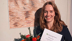 Porträt von Lea Hornscheidt. Sie steht vor ihrem Gewinnerbild. Sie hält Blumen und die Urkunde in der Hand.