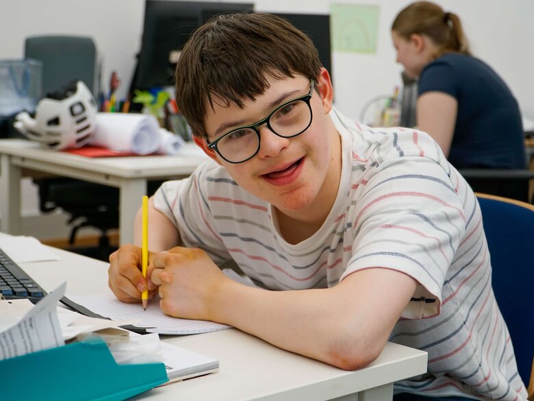 Lukas sitzt an einem Scheibisch in einem der AW-ZIB - Büros. Er hält einen Stift in der Hand und lächelt in die Kamera.