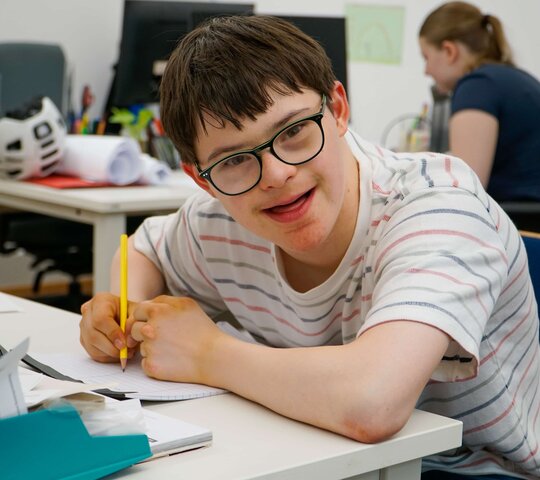 Lukas sitzt an einem Scheibisch in einem der AW-ZIB - Büros. Er hält einen Stift in der Hand und lächelt in die Kamera.