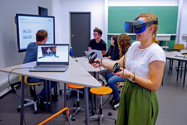 Ein Beispiel digitaler Lehre: Eine Studentin der Pädagogischen Hochschule Heidelberg trägt eine VR-Brille und hält die Controller in der Hand. Im Hintergrund sitzt eine Gruppe zusammen am Stehtisch und diskutiert vor einem Whiteboard.