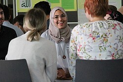 Das Bild zeigt mehrere Personen auf Stühlen sitzend. Eine Frau hat eine muslimische Kopfbedeckung an.