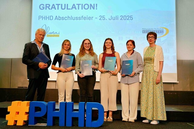 Zu sehen sind Alumni, die auf einer Bühne stehen und ihre Bestehensbriefe in PHHD-Mappen hochhalten. Gemeinsam mit Rektorin Vach & Prorektor Dammer (a.D.), welche ihnen die Mappen überreicht haben.