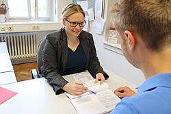 Frau die einem Mann gegenüber sitzt und ihm etwas auf einem Blatt erklärt.