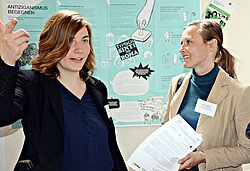 Das Symbolbild zeigt zwei Frauen im Gespräch vor einem Plakat mit Informationen zu "Antiziganismus begegnen". Copyright Pädagogische Hochschule Heidelberg.