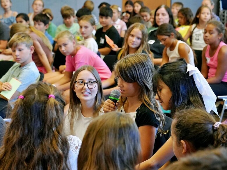 Eine Studentin in Mitten von Grundschüler:innen. Eine der Schülerinnen spricht aktiv in ein Mikrofon. Aufgenommen wurde das Foto in der Festhalle der Pädagogischen Hochschule Heidelberg.
