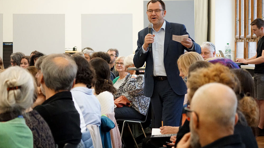 Frank Ziegele geht mit dem Mikro durch die vollbesetzte Aula. Er spricht deutlich mit den Händen.